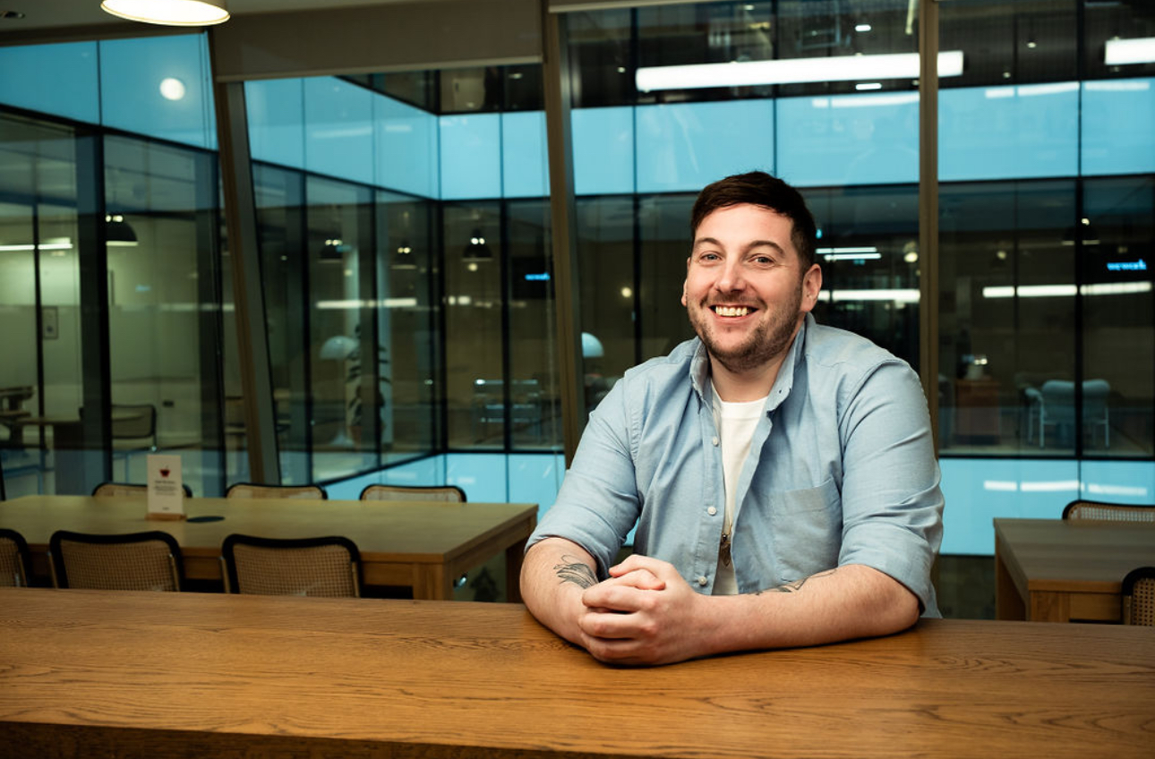 picture of smiling man with beard sat behind a desk infront of windows